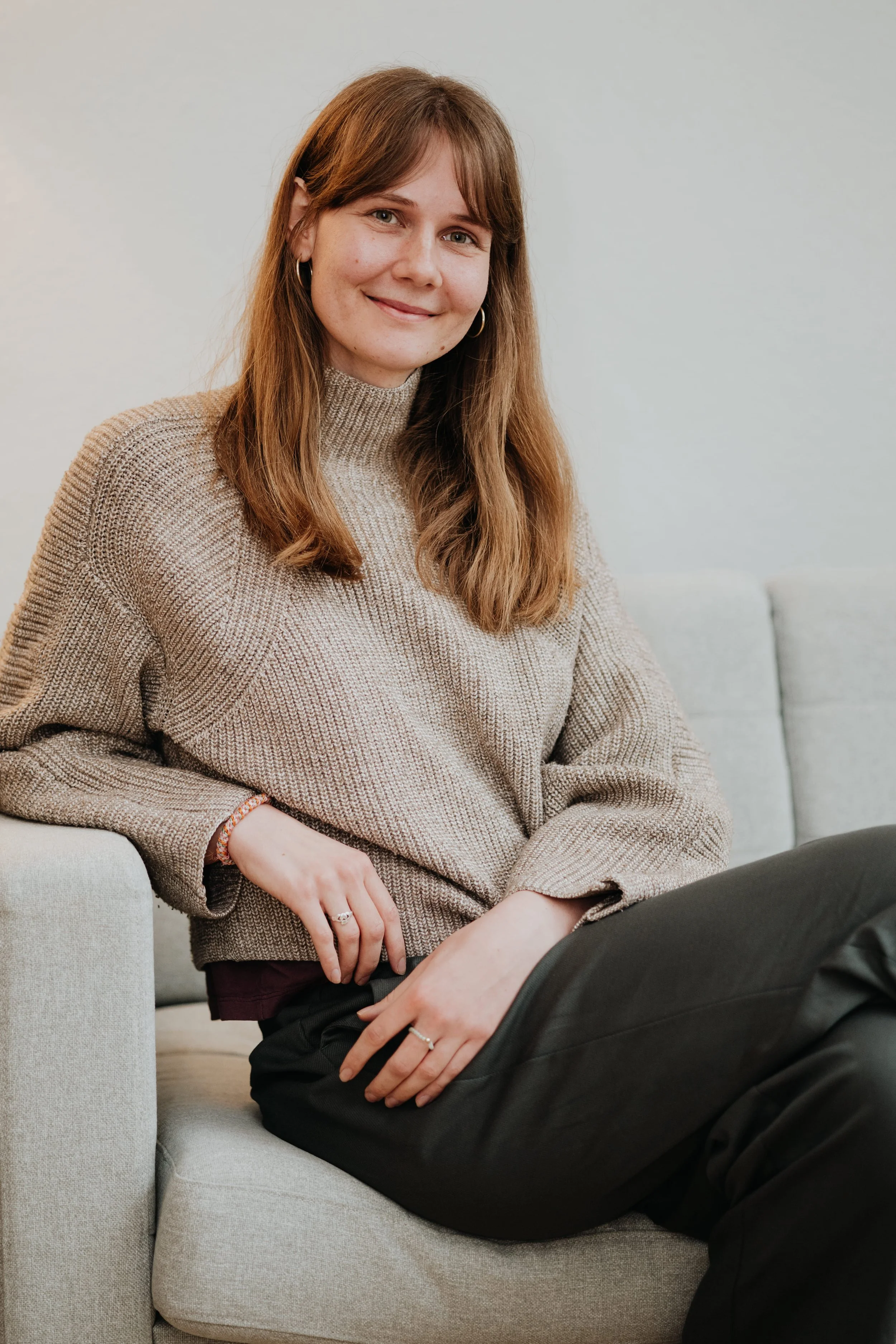 Eine junge Frau mit braunen Haaren sitzt entspannt auf einem hellen Sofa vor einem weißen Hintergrund, trägt einen beigen Pullover und lächelt freundlich in die Kamera.