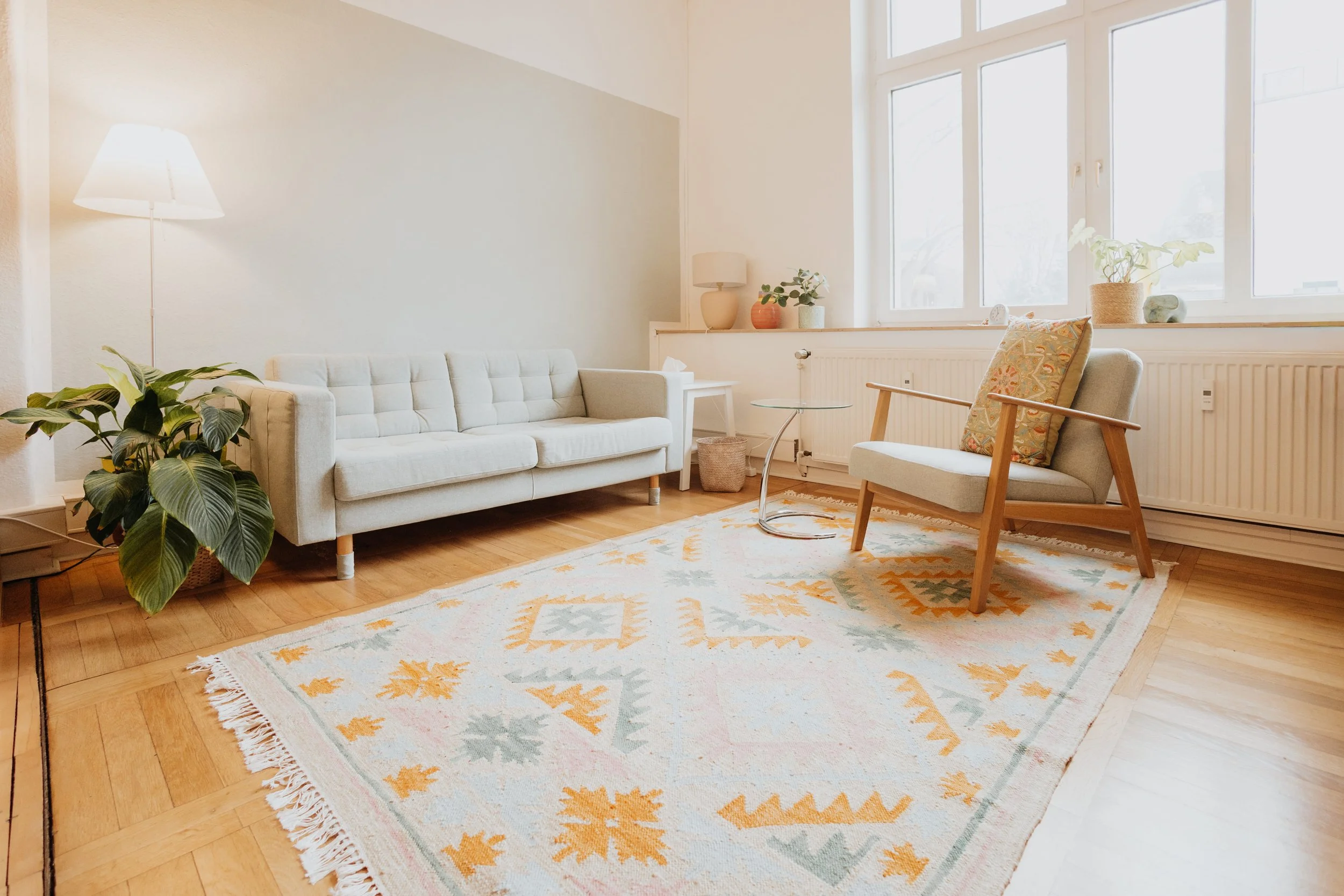 Helles Wohnzimmer mit weißem Sofa, Sessel, Teppich, Pflanzen und großen Fenstern