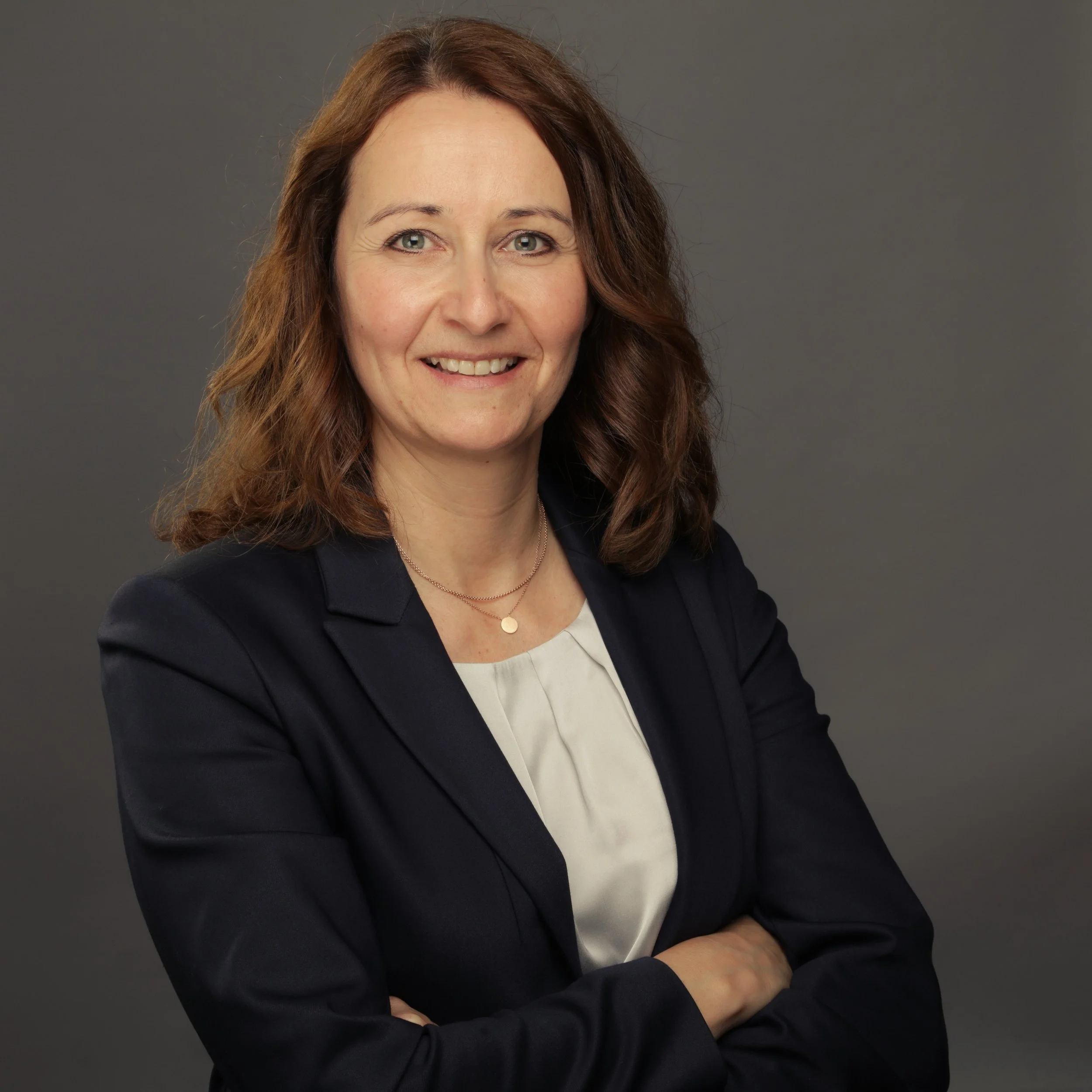 Porträt einer Frau mit braunen, mittellangen Haaren, in dunkler Blazer Jacke und weißem Hemd, vor grauem Hintergrund.