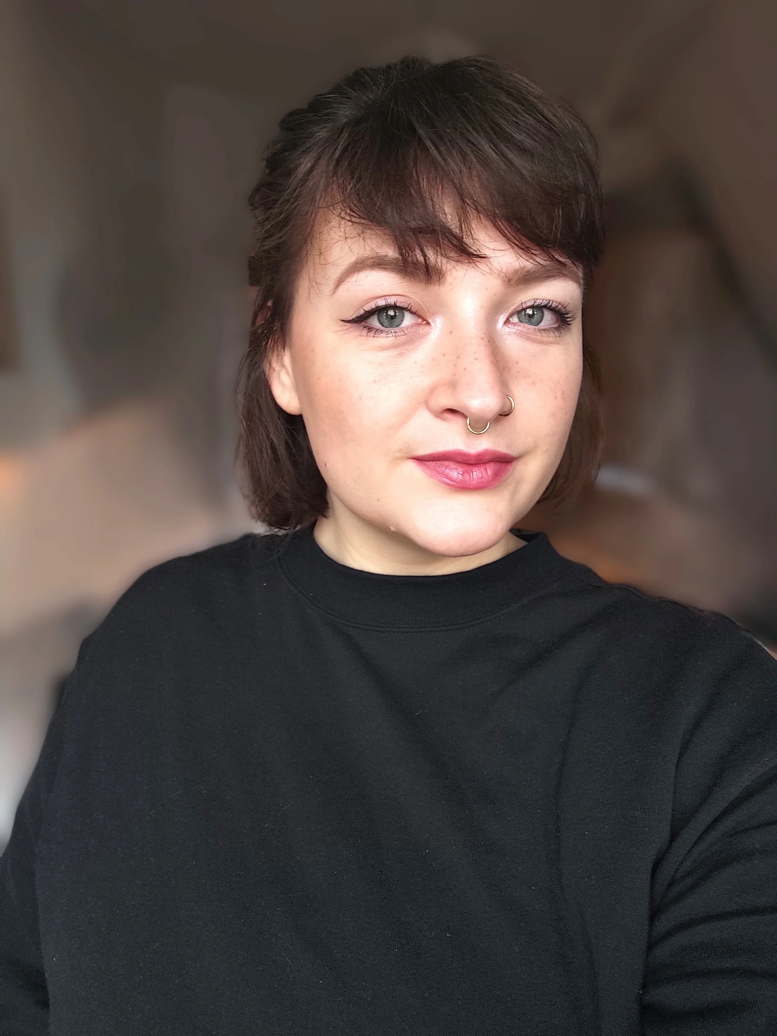 Nahaufnahme eines jungen Mädchens mit kurzen braunen Haaren, Nasenpiercing und schwarzen Pullover, vor einem unscharfen Hintergrund.