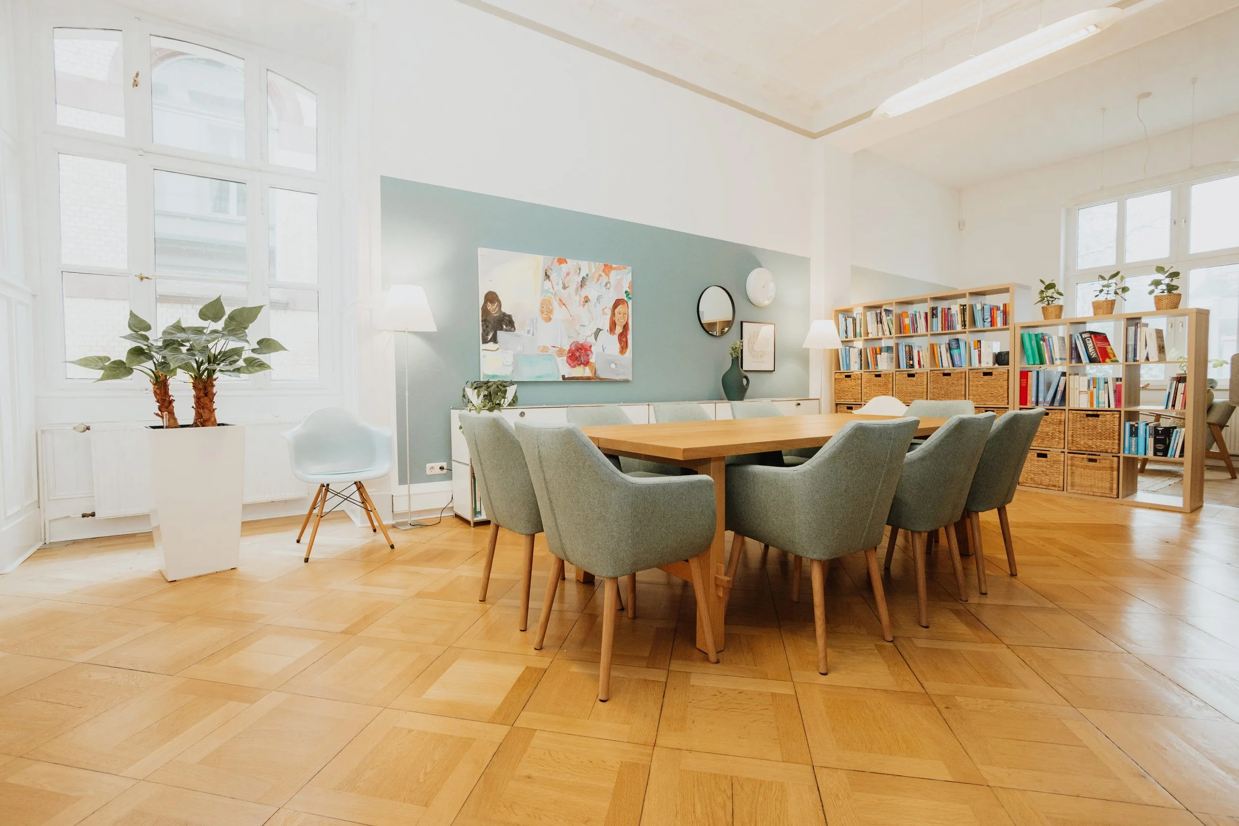 Heller, moderner Esszimmer- oder Gemeinschaftsraum mit Holztisch, grauen Stühlen, Bücherregalen, Pflanzen und Kunstdekorationen, viel Tageslicht durch große Fenster.