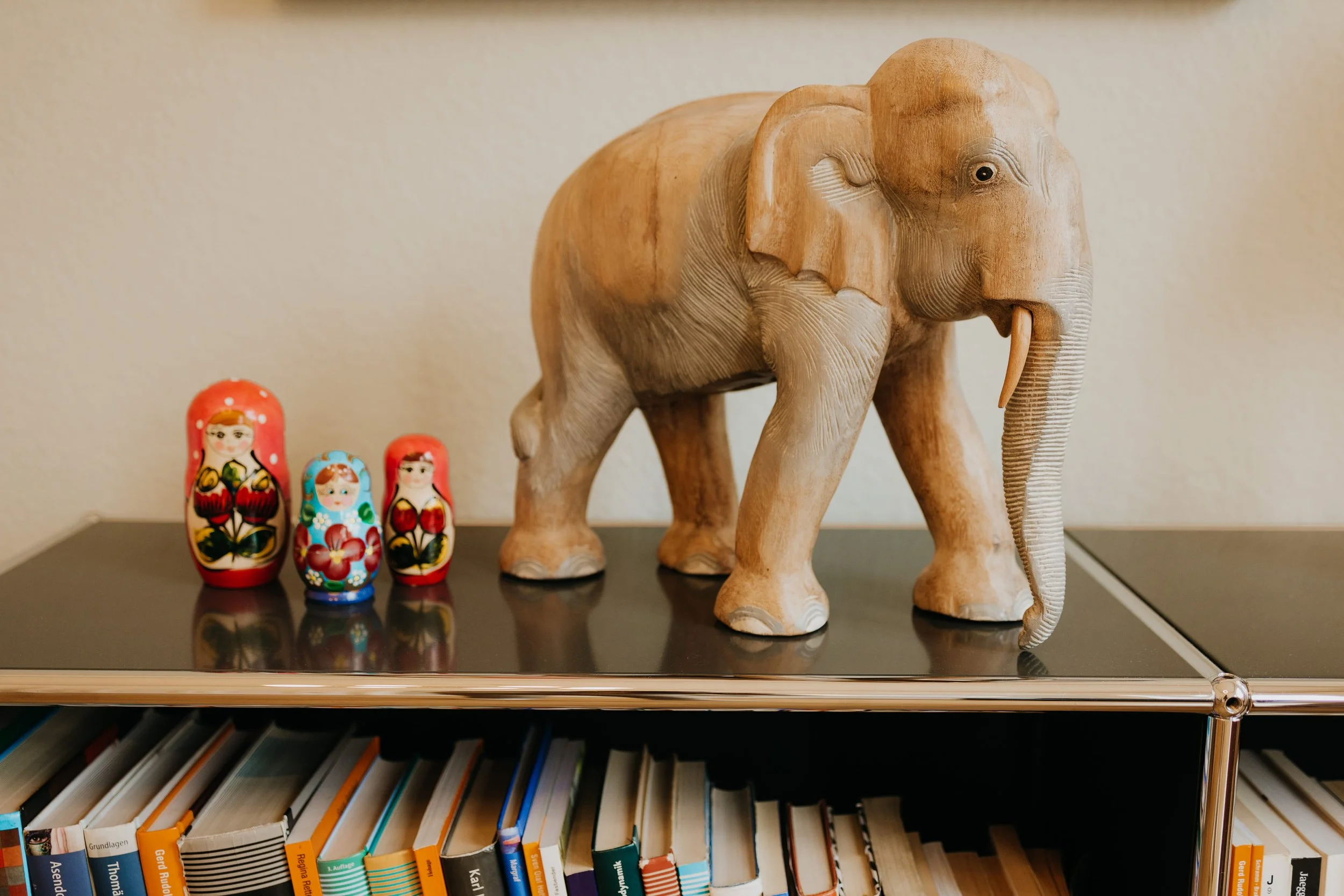 Deko-Set mit einem großen aus Holz geschnitzten Elefanten und drei bemalten russischen Matroschka-Puppen auf einem schwarzen Tisch vor einem Bücherregal.