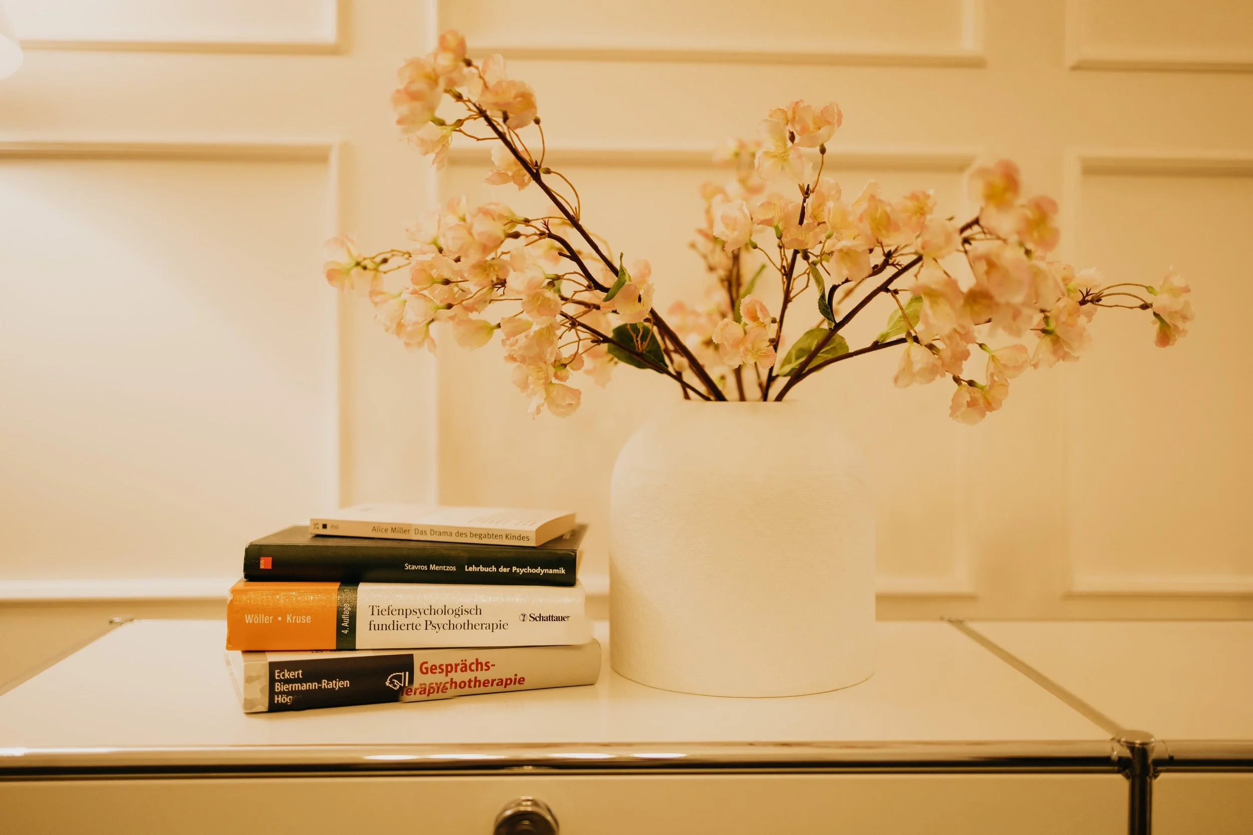 Weiße Vase mit rosa Blüten neben aufgestapelten Büchern auf weißem Möbelstück.