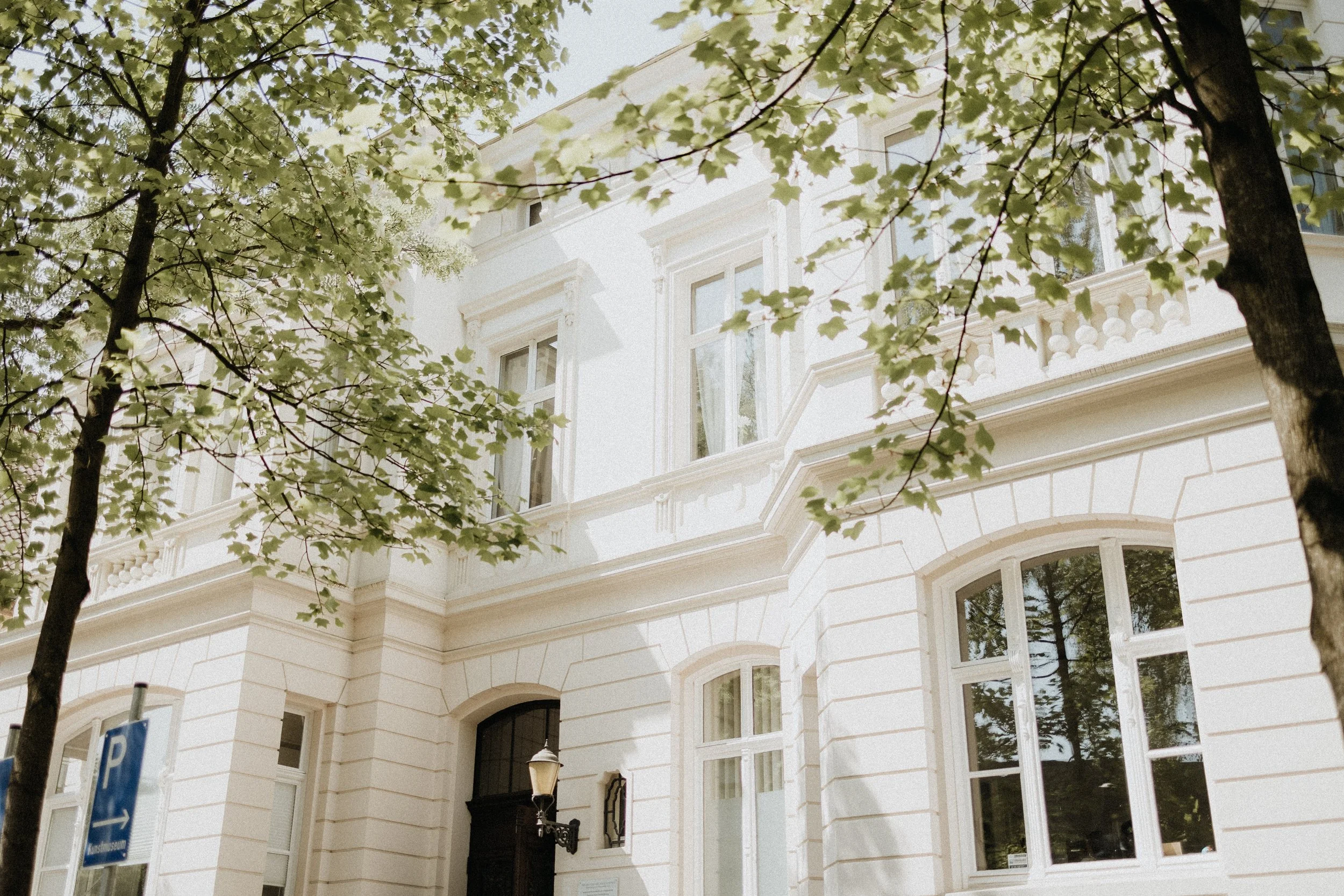Ein weißes, historisches Gebäude mit mehreren Fenstern und einer alten Laterne an der Tür, umgeben von grünen Blättern. Es scheint ein sonniger Tag zu sein.