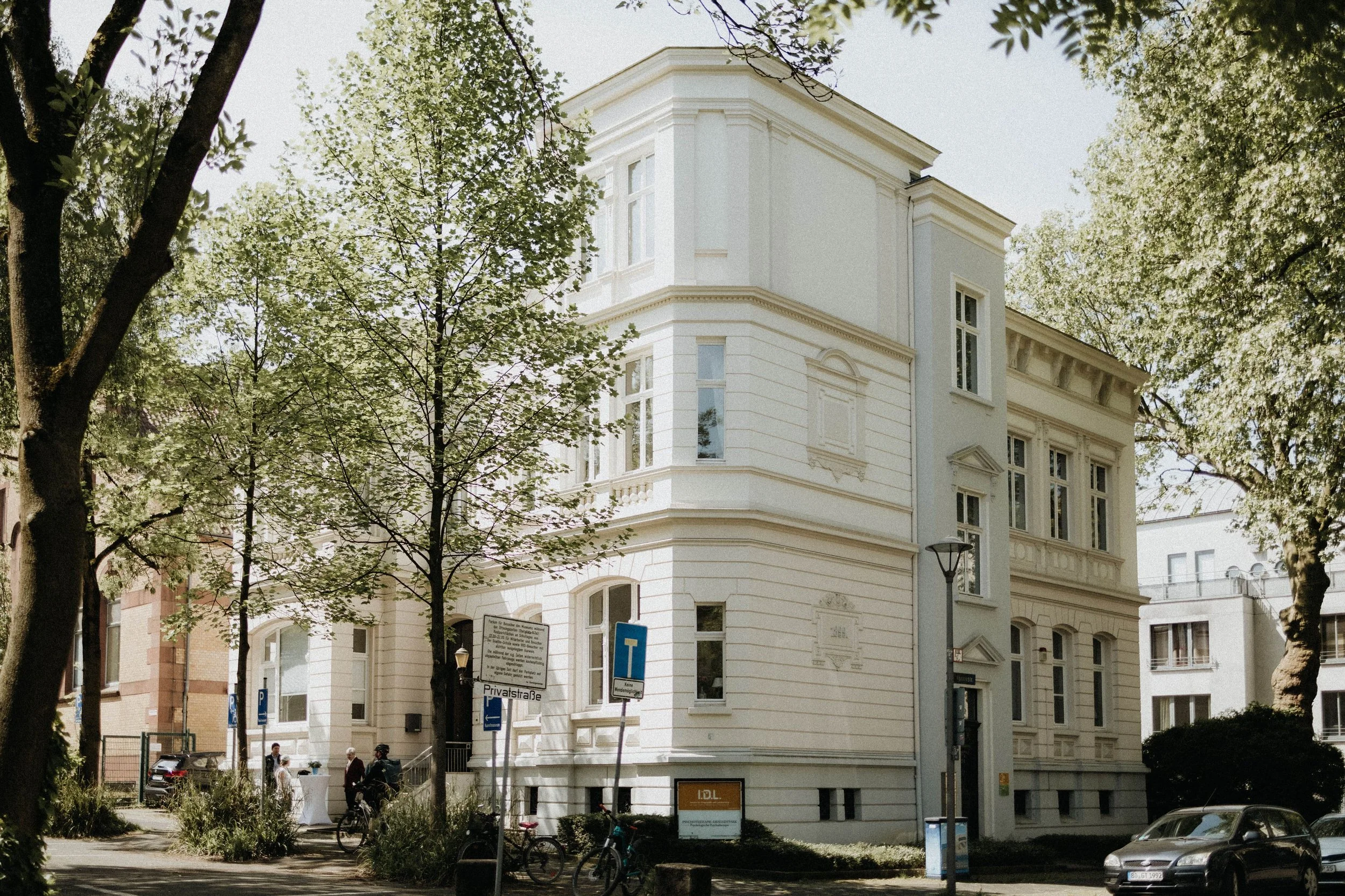 Ein weißes, mehrstöckiges Wohnhaus mit klassischen architektonischen Elementen, umgeben von Bäumen und Autos, auf einer ruhigen Straße in einer Stadt.