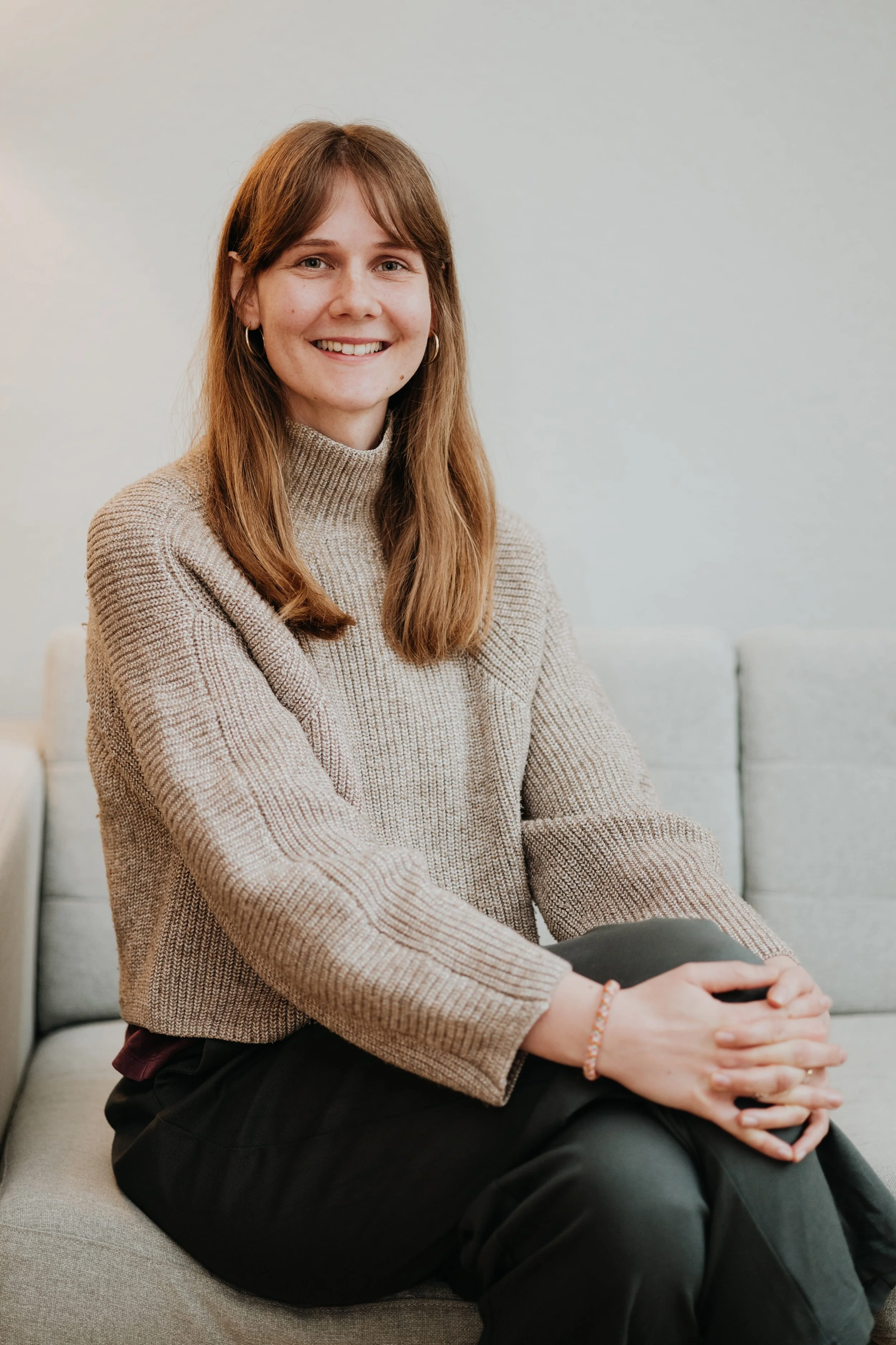 Junge Frau mit braunen Haaren, grauem Pullover und schwarzen Hosen sitzt auf einem Sofa in einem hellen Raum.