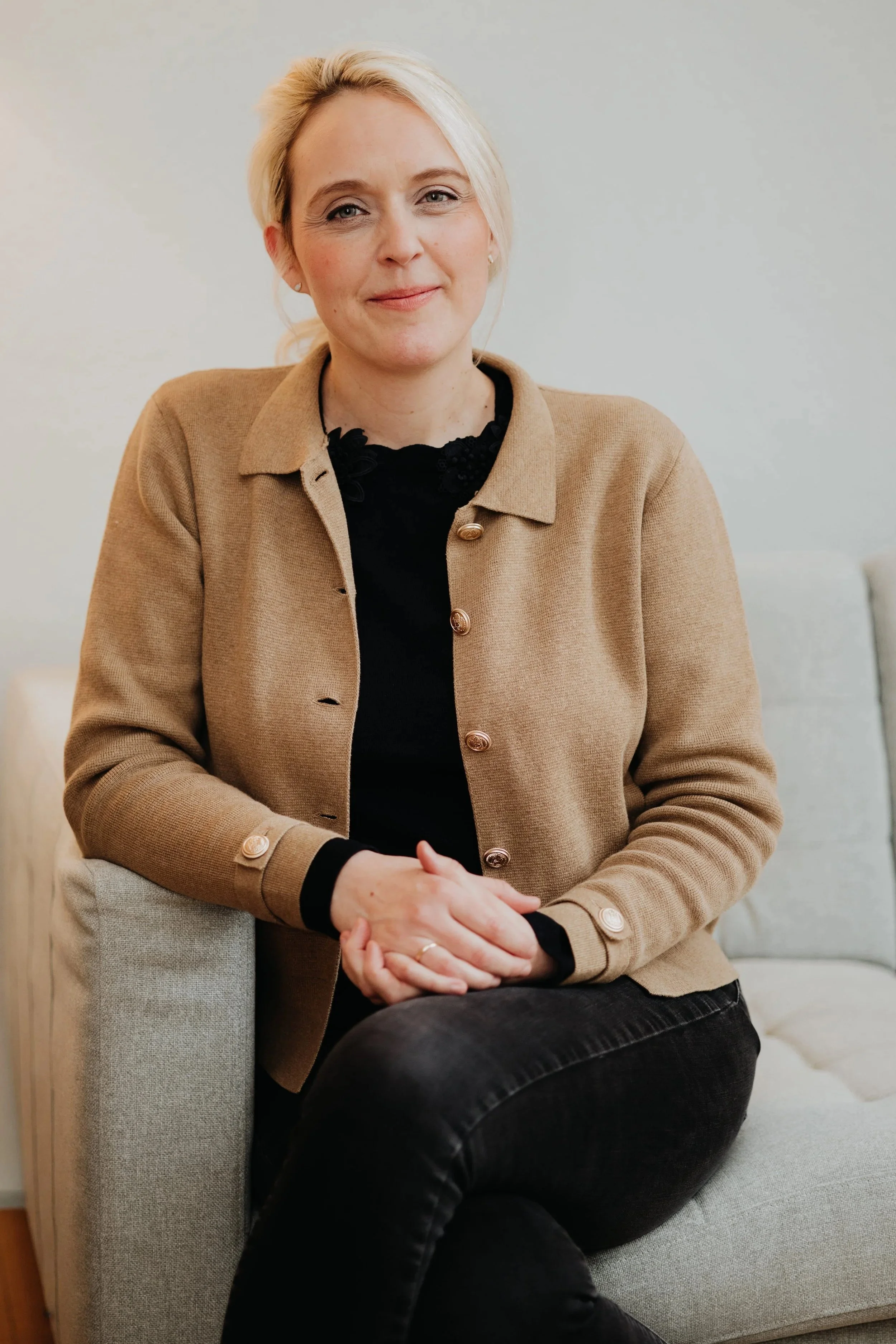 Eine Frau sitzt entspannt auf einem Sofa, trägt eine beige Jacke und schwarze Hose, hat blonde Haare und lächelt leicht.